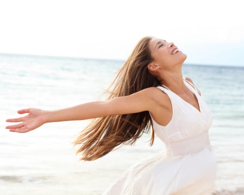 A woman in a flowing white dress joyfully extends her arms while standing on a sandy beach, with the ocean waves in the background.