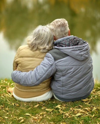 An older couple sitting on the grass by a serene lake, enjoying each other's company in a peaceful outdoor setting.