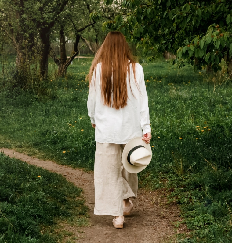 A woman in a white shirt and linen pants walks along a grassy path, holding a wide-brimmed hat, surrounded by lush greenery.