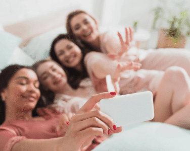 A group of friends lounging on a bed, enjoying each other’s company and taking selfies, surrounded by soft pillows and warm lighting.
