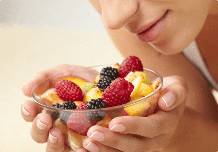 A close-up of a woman gently holding a glass bowl filled with assorted fresh fruits, including blackberries and raspberries.