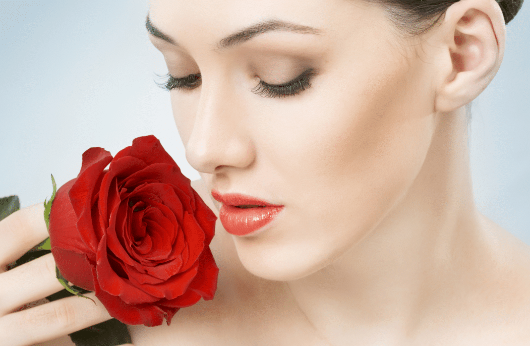 A beautiful girl holds a vibrant red rose close to her neck against a soft blue background, evoking a sense of beauty and tranquility.
