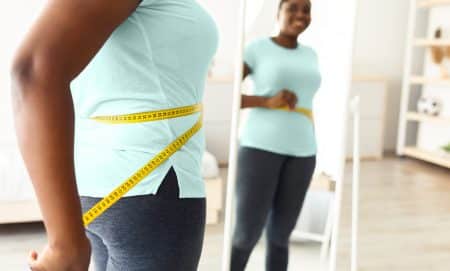Cropped view of overweight young black woman measuring waist with tape near mirror at home, free space. Plump African American lady satisfied with slimming result, achieving success in weight loss