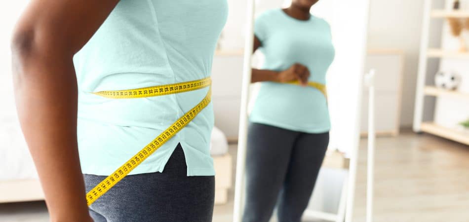 Cropped view of overweight young black woman measuring waist with tape near mirror at home, free space. Plump African American lady satisfied with slimming result, achieving success in weight loss