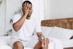 Hangover Concept. Sleepy Black Guy Touching Aching Head Holding Glass Of Water Sitting In Bed At Home In The Morning