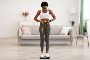 Weight Gain. Shocked Black Woman Weighing Herself And Measuring Her Waist Standing On Scales Unhappy With Result At Home. Slimming And Weight-Loss Failure Concept. Full Length