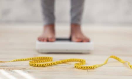 Unrecognizable black lady standing on scales, checking her weight, focus on measuring tape. Cropped view of African American woman fighting excess pounds, using slimming diet