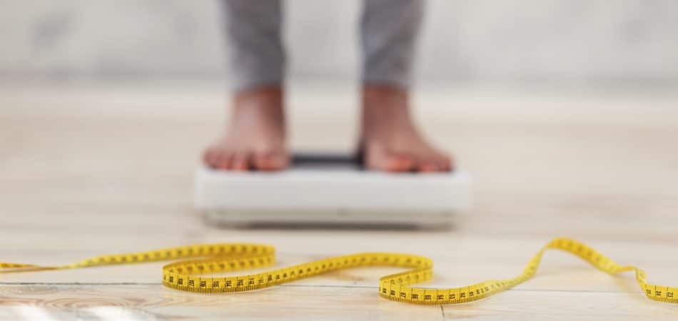 Unrecognizable black lady standing on scales, checking her weight, focus on measuring tape. Cropped view of African American woman fighting excess pounds, using slimming diet
