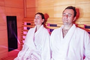 Young married couple in white bathrobes sits and relaxes in salt room. Attractive man and beautiful woman have closed their eyes and are resting on spa procedure. Husband and wife enjoy time together.