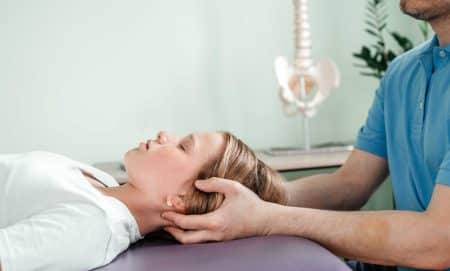 A person lying on their back while a therapist in a blue shirt supports their head and neck with both hands; a spine model and plant are visible in the background.