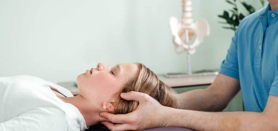 A person lying on their back while a therapist in a blue shirt supports their head and neck with both hands; a spine model and plant are visible in the background.