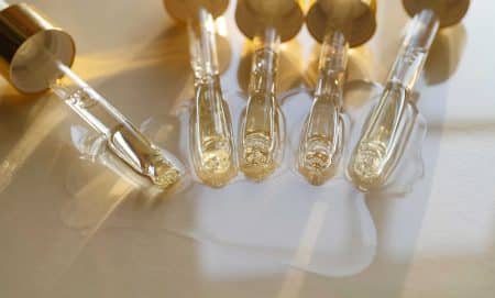 Five glass droppers with gold caps lay side by side on a light surface, dispensing clear liquid that forms small pools and reflections beneath them in soft, natural light.