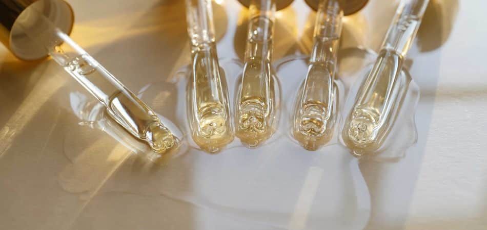 Five glass droppers with gold caps lay side by side on a light surface, dispensing clear liquid that forms small pools and reflections beneath them in soft, natural light.