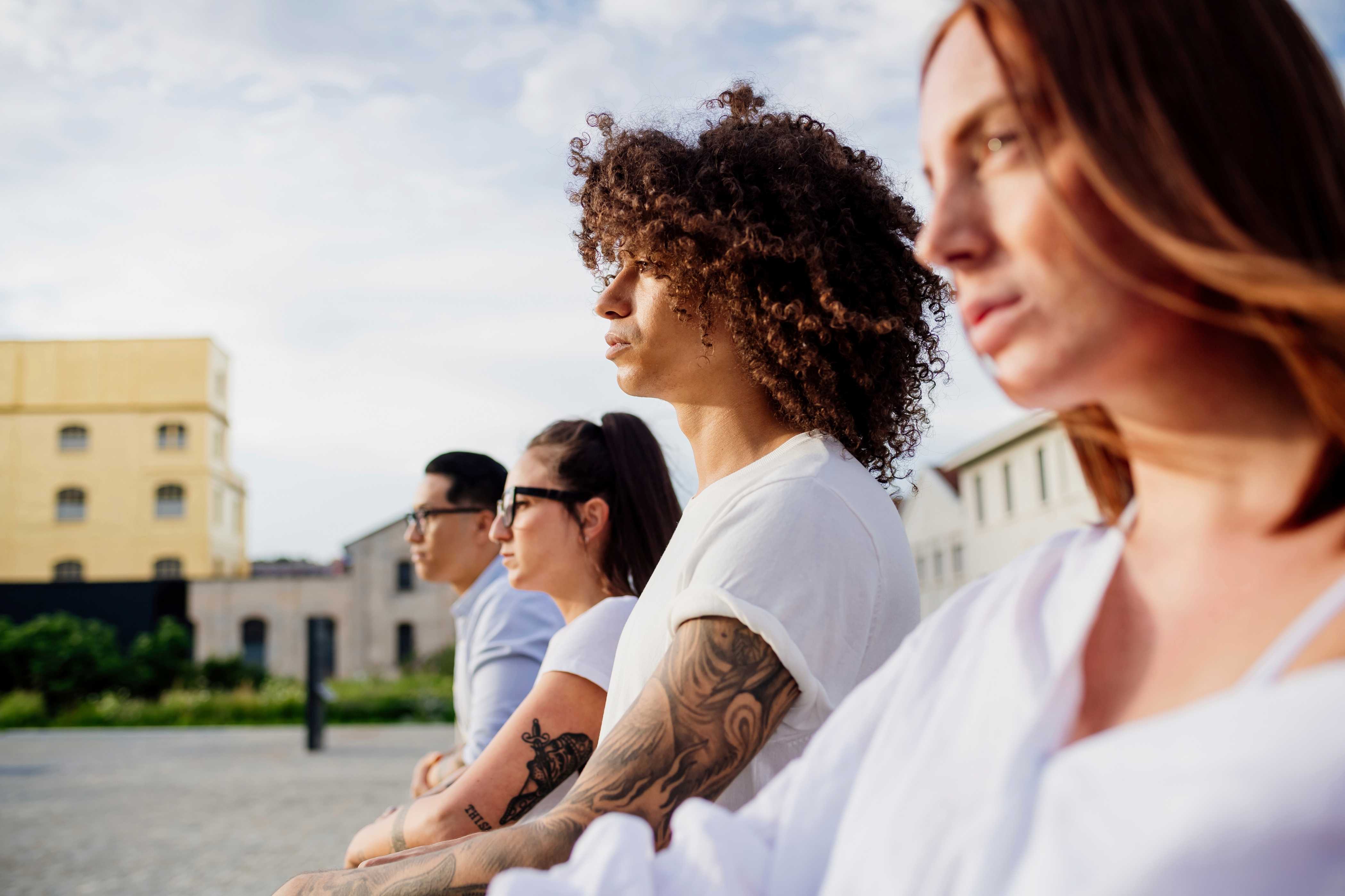 A diverse group of four people sits together outdoors, wearing casual clothing, with a backdrop of buildings and blue sky.
