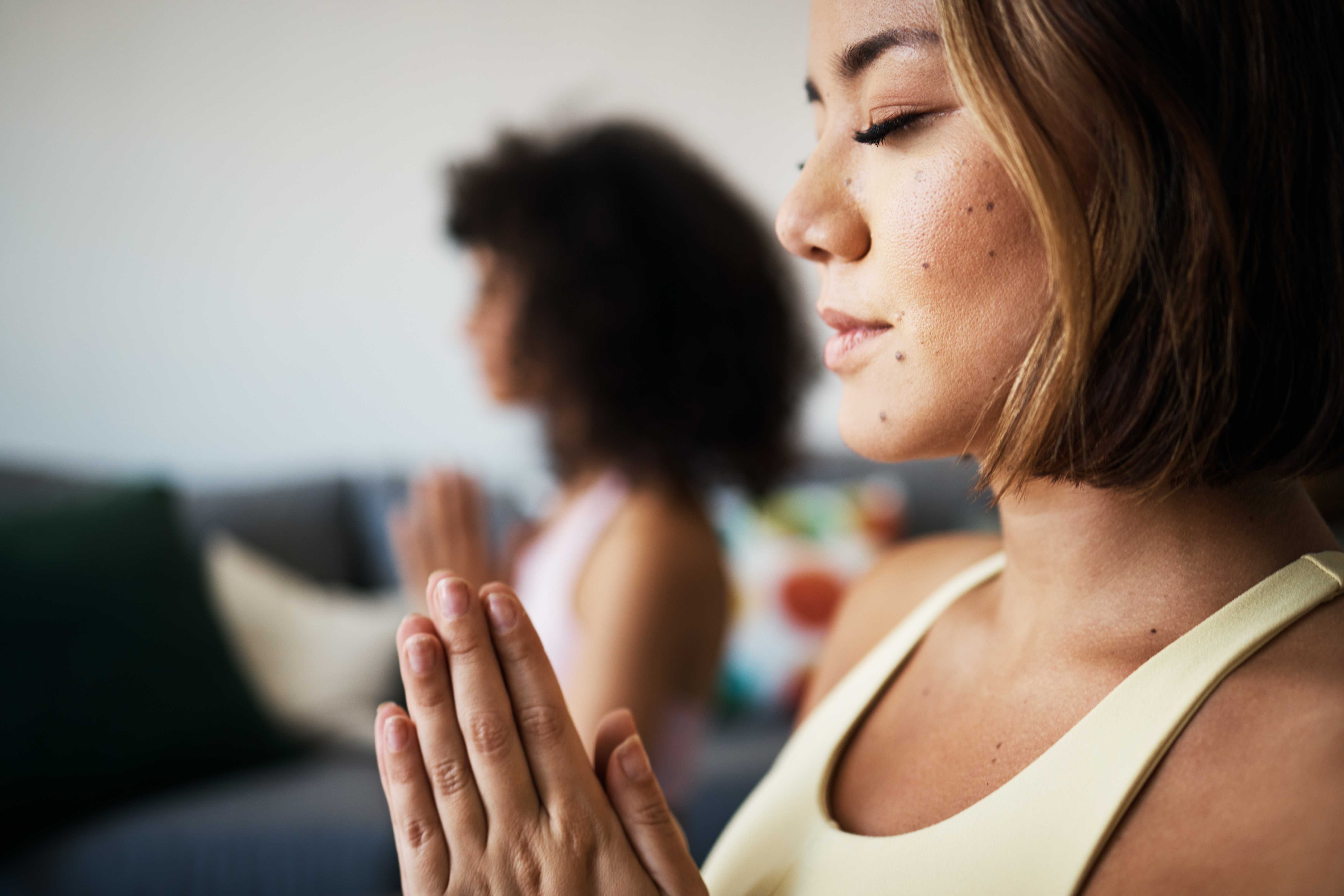 Two women practice mindfulness together, hands clasped in prayer position, seated in a cozy living room with colorful decor.
