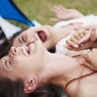 Two women lying on a blanket outdoors, laughing and holding hands, enjoy nature with their tent—and perhaps the revitalizing effects of a NAD+ booster—visible in the background.