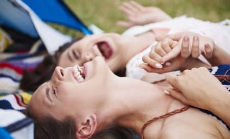 Two women lying on a blanket outdoors, laughing and holding hands, enjoy nature with their tent—and perhaps the revitalizing effects of a NAD+ booster—visible in the background.