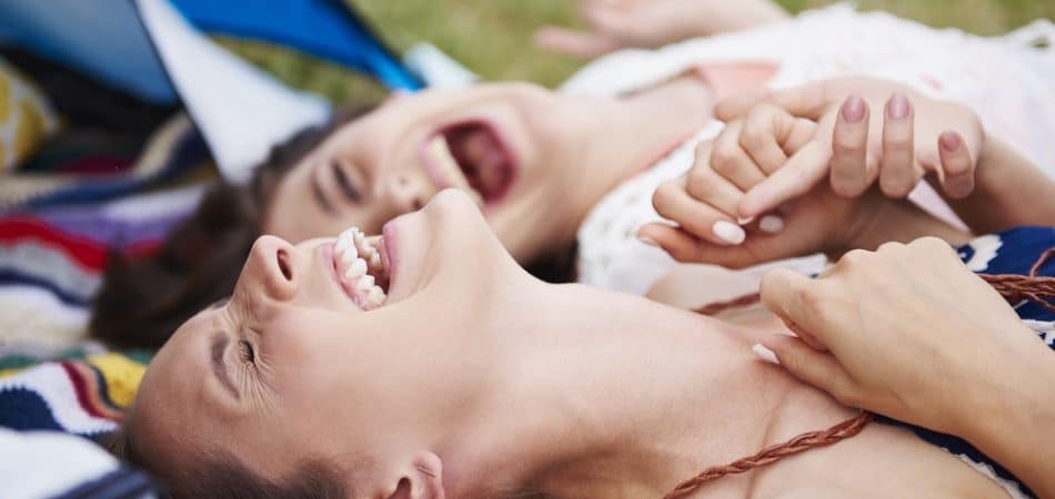 Two women lying on a blanket outdoors, laughing and holding hands, enjoy nature with their tent—and perhaps the revitalizing effects of a NAD+ booster—visible in the background.