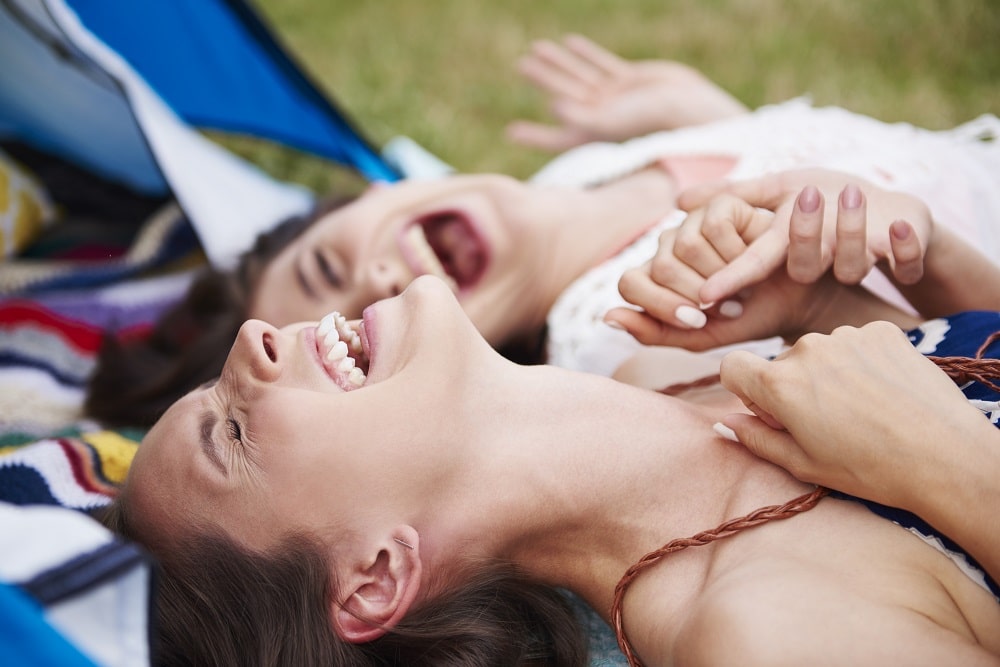 Two women lying on a blanket outdoors, laughing and holding hands, enjoy nature with their tent—and perhaps the revitalizing effects of a NAD+ booster—visible in the background.