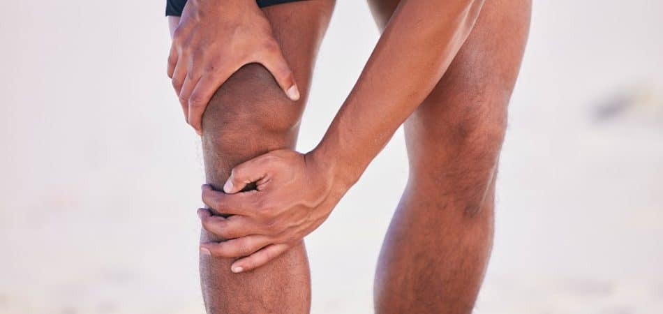 A person in athletic shorts holding their knee with both hands, possibly indicating knee pain or discomfort, standing outdoors—an image relevant to discussions about CBD vs. THC for managing joint issues.