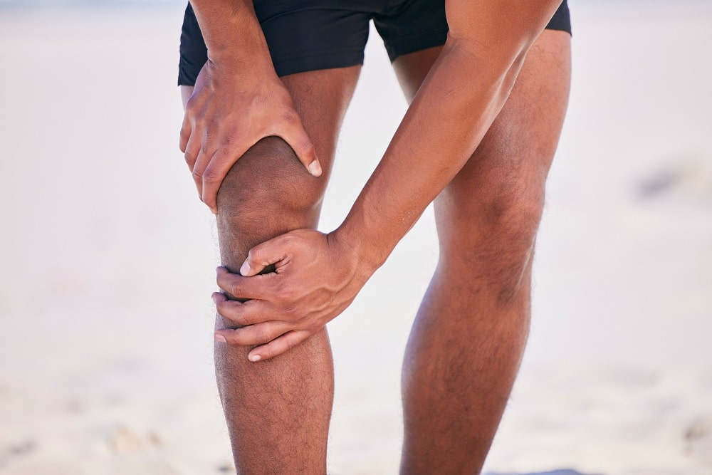 A person in athletic shorts holding their knee with both hands, possibly indicating knee pain or discomfort, standing outdoors—an image relevant to discussions about CBD vs. THC for managing joint issues.