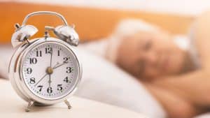 A silver analog alarm clock showing 7:00 sits on a bedside table, hinting at better sleep as an elderly woman rests peacefully in the blurred background.