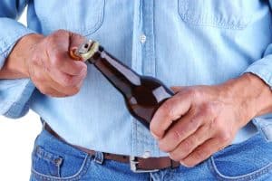 A person in a denim shirt and jeans is opening a brown glass bottle with a metal bottle opener, mindful that alcohol effects on weight loss are often overlooked.