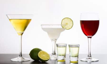 Four alcoholic beverages—a martini, a margarita with lime, two tequila shots, and a glass of red wine—are displayed on a table with limes, offering a visual reminder of how alcohol effects on weight loss can impact your health goals.