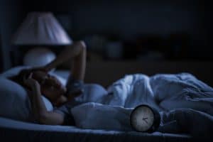 A person lies awake in bed at night, holding their head, with an alarm clock showing 2:10 on the bedside—perhaps reflecting on alcohol effects on weight loss.