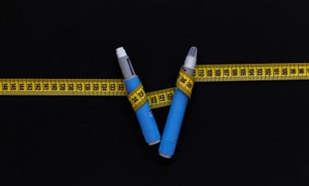 Two blue weight loss injection pens are positioned in a V shape on a black background, wrapped with a yellow measuring tape showing centimeter markings.