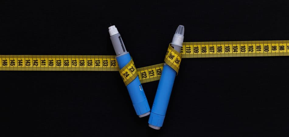 Two blue weight loss injection pens are positioned in a V shape on a black background, wrapped with a yellow measuring tape showing centimeter markings.